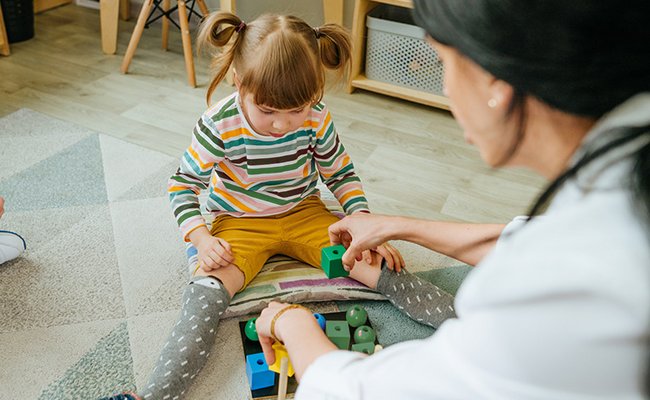 Mädchen sitzt auf dem Boden und sortiert mit Erzieherin Bausteine