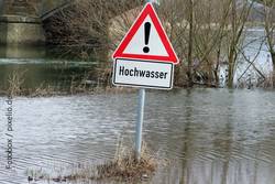 Foto: Dreieckiges Warnschild, rot umrandet, mit der Aufschrift "Hochwasser", inmitten einer gefluteten Naturlandschaft